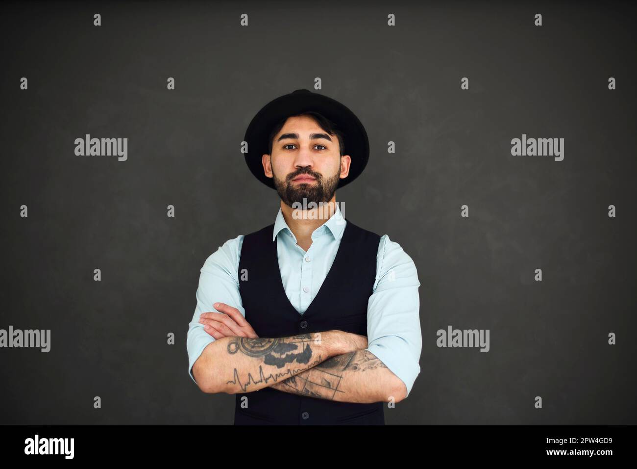 Studio portrait of handsome ethnic male with crossed tattooed arms, dressed in a blue shirt and black hat. Young stylish man on dark background Stock Photo