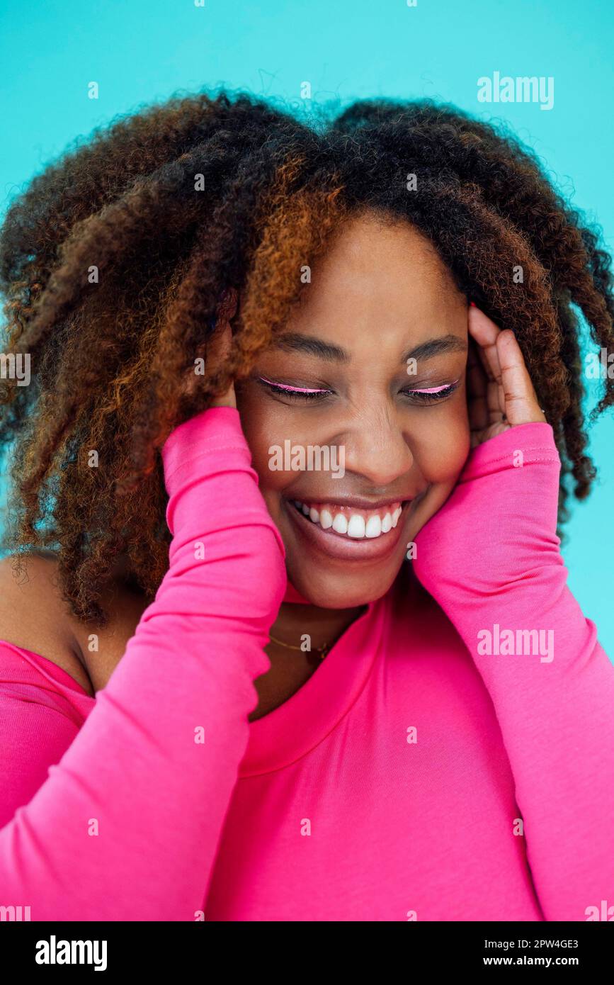 Dreamy young beautiful African American woman with bright eyelines wearing pink bodysuit over blue wall keeps hands pressed together under chin Stock Photo