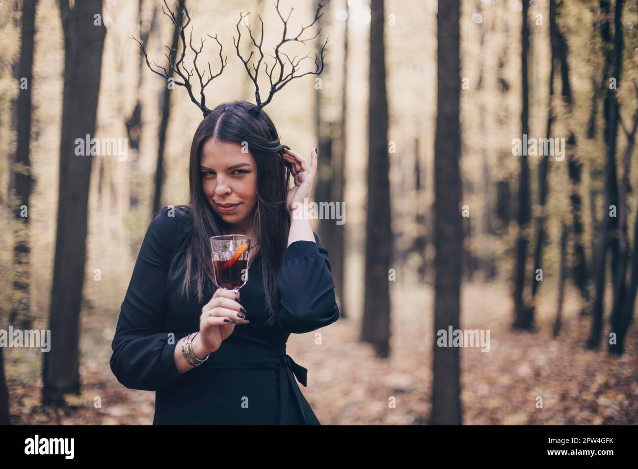 Mysterious woman in black dress wearing gothic deer antlers headband holding mulled wine in glass while standing in autumn forest, looking at camera Stock Photo