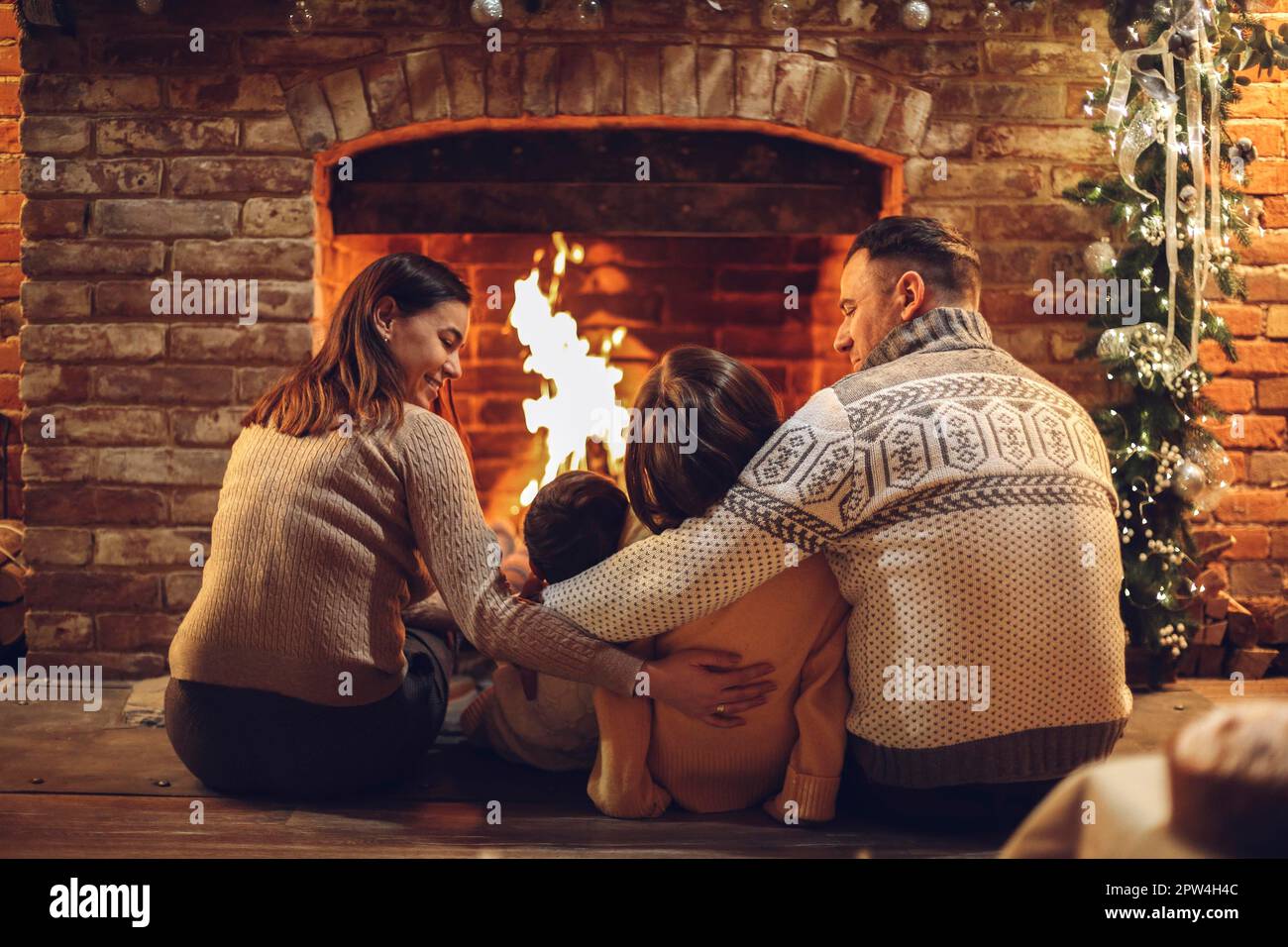 Happy young family parents with two small kids gathering around brick fireplace in cozy and warm country house during Christmas holidays in winter. Stock Photo