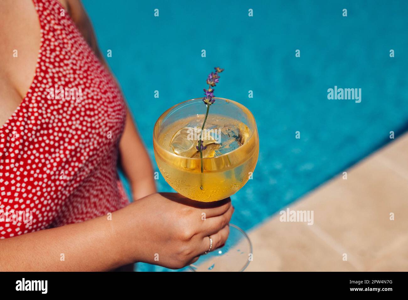 Young woman hand with red manicure holds a glass of champagne. Wine cocktail with lemon wedges. Sunny day and reflection of rays in blue water on Stock Photo