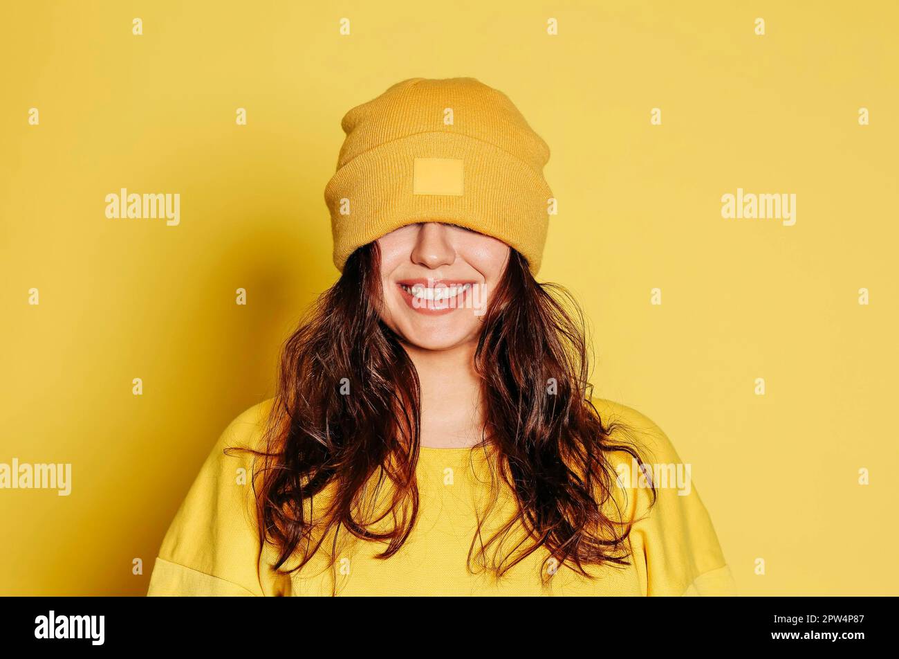 Happy young female in trendy yellow sweatshirt and knitted hat covering eyes smiling brightly against yellow background Stock Photo