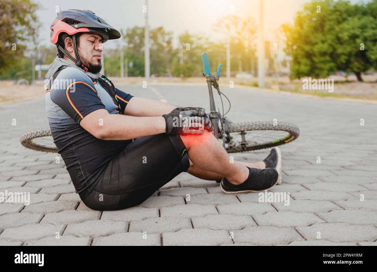 Cyclist with knee pain outdoors. Male cyclist sitting on the pavement with knee pain. Concept of a cyclist with knee injury outdoors Stock Photo