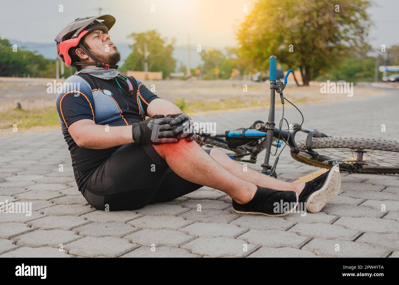 Male cyclist sitting on the pavement with knee pain. Concept of a cyclist with knee injury outdoors, Cyclist with knee pain outdoors Stock Photo