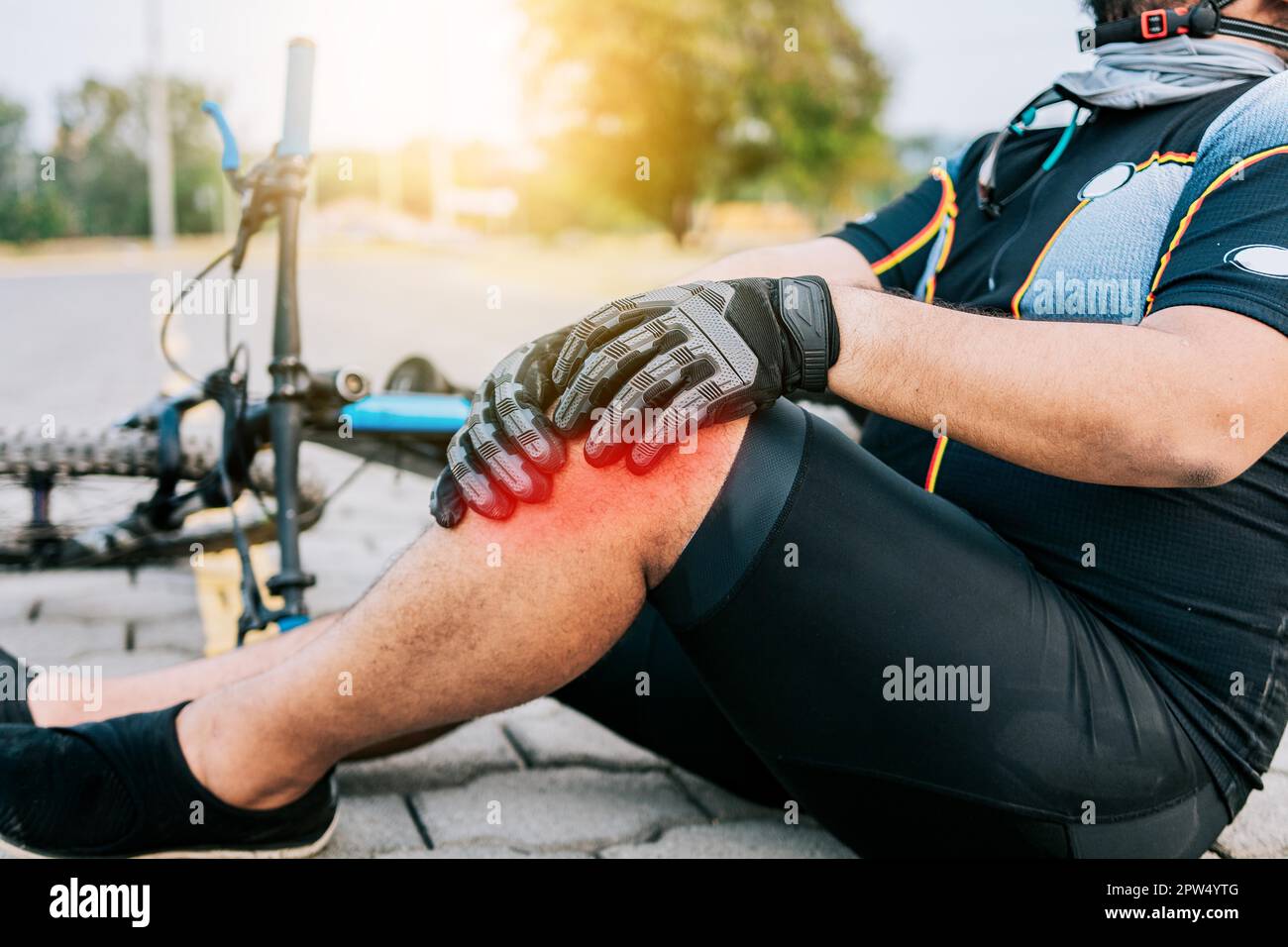 Concept of cyclist with knee injury outdoors. Cyclist with knee pain outdoors. Male cyclist sitting on the pavement with knee pain Stock Photo
