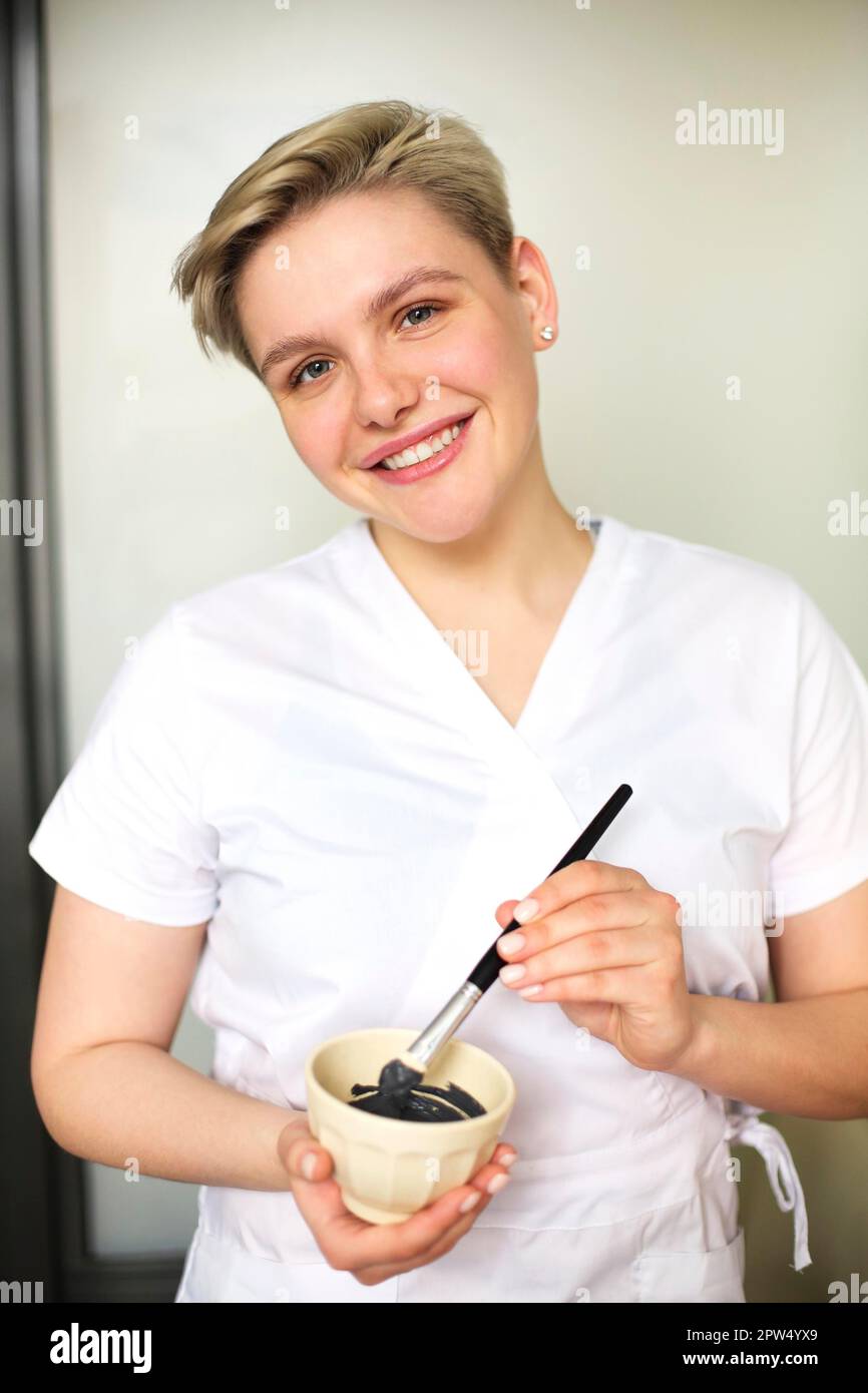 Content female beautician preparing clay facial mask for skin care treatment in beauty salon and looking at camera Stock Photo