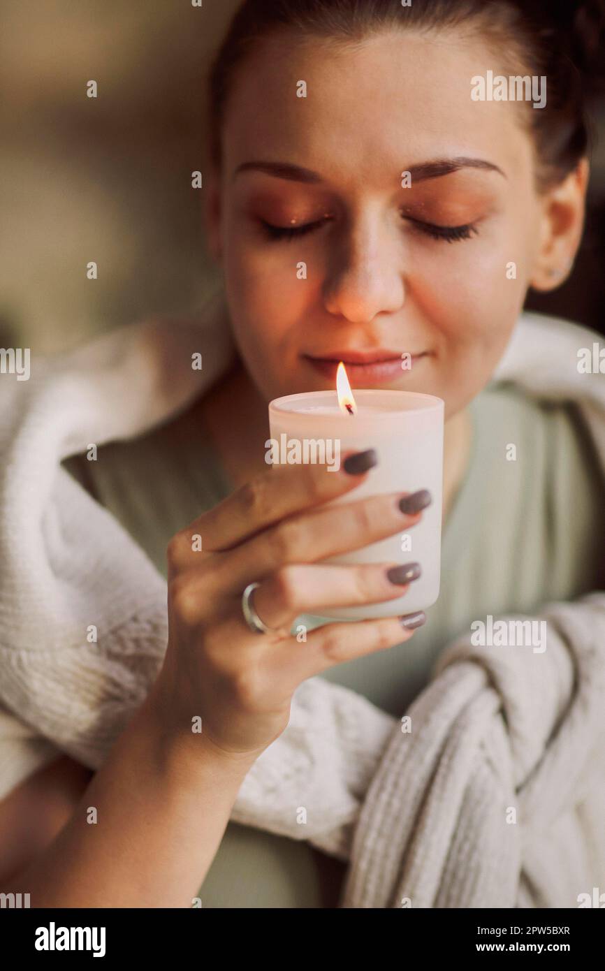Side portrait of young calm woman holding lit candle in hands, closing eyes bringing close to
