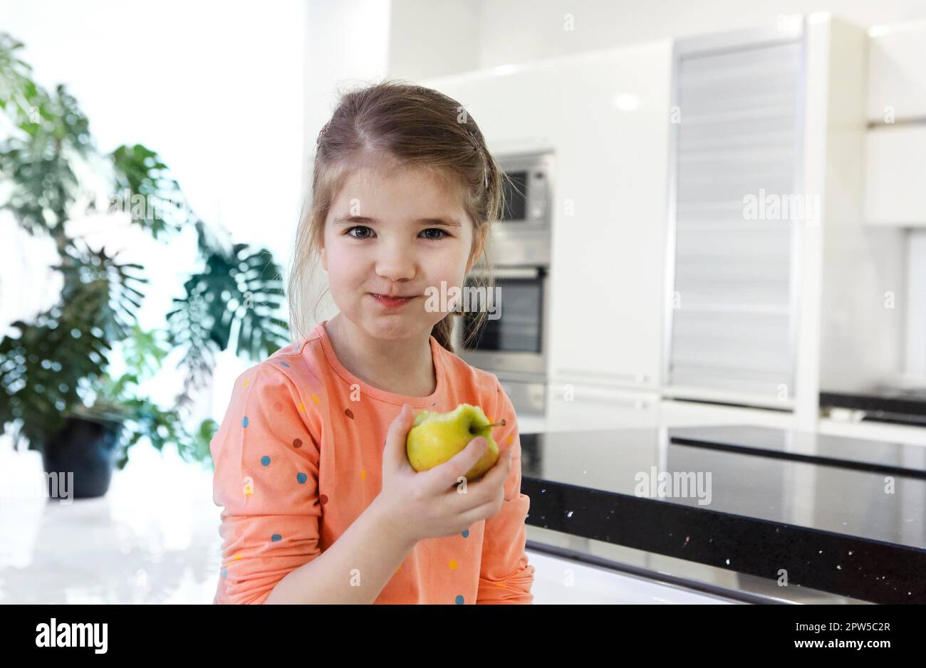 Little caucasian girl eats a big red apple in the kitchen. Beautiful little girl bites and eats an apple. Looks to the camera holding fruit. Close up Stock Photo