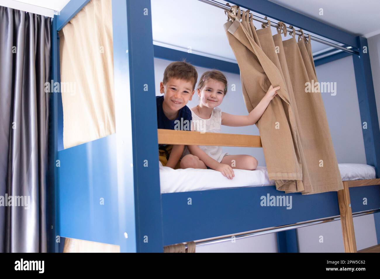 Caucasian little sibling are sitting on the bunk bed and looking to camera with big smile