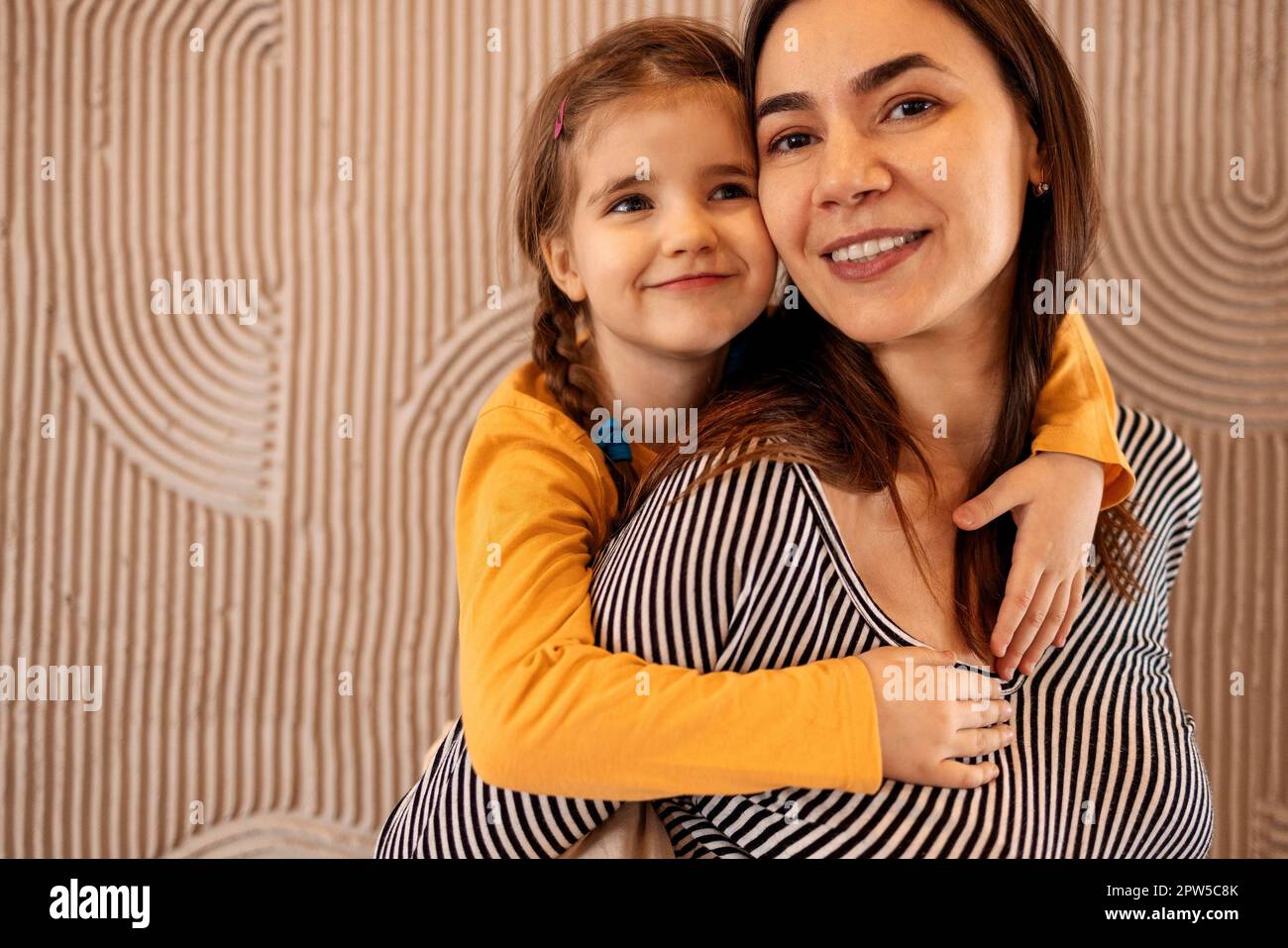 Mom and little daughter hug on beige background with copy space, happy family, parenthood. Concept of maternal love and care Stock Photo