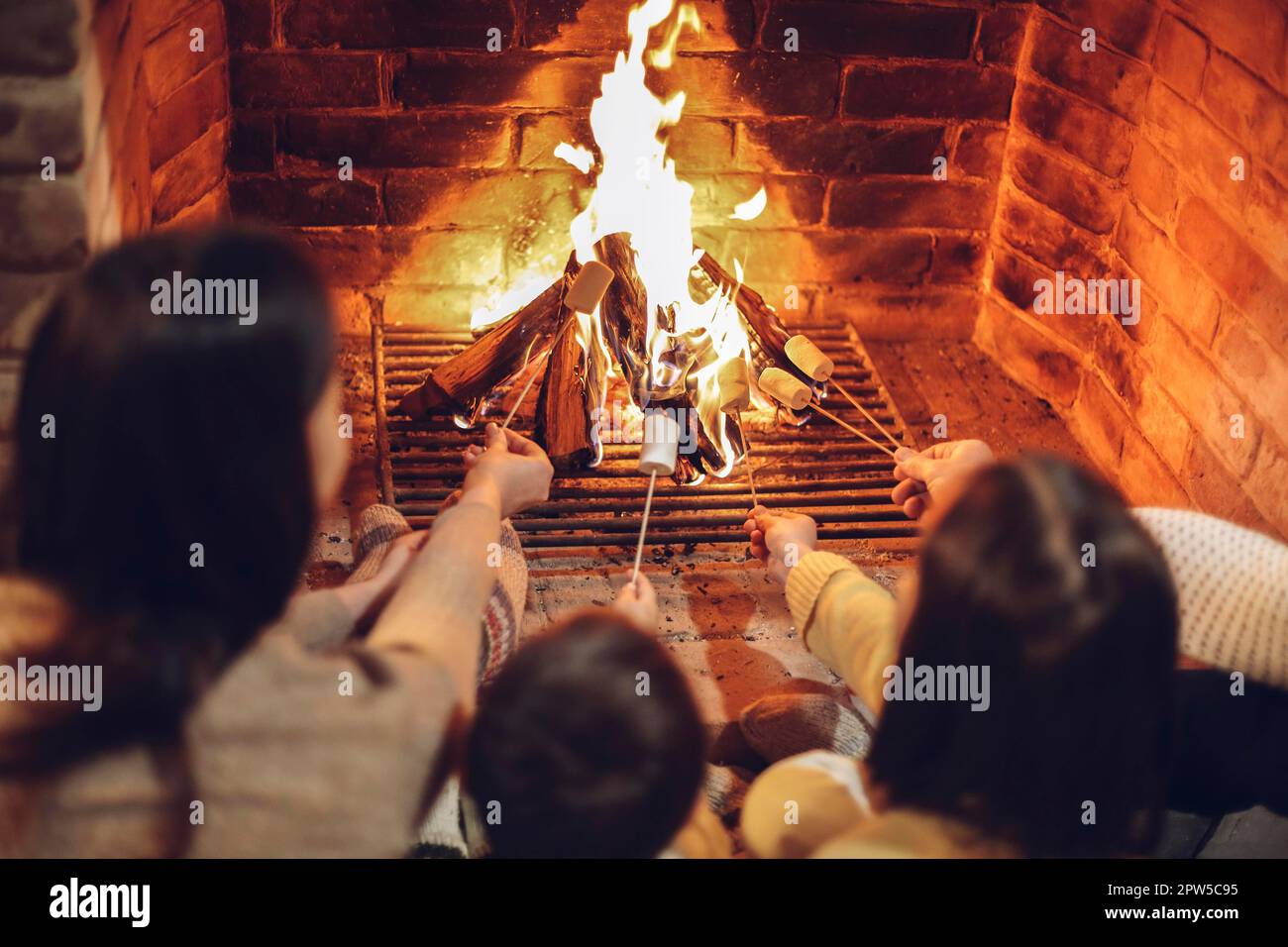 Happy young family parents with two small kids gathering around brick fireplace in cozy and warm country house during Christmas holidays in winter. Stock Photo