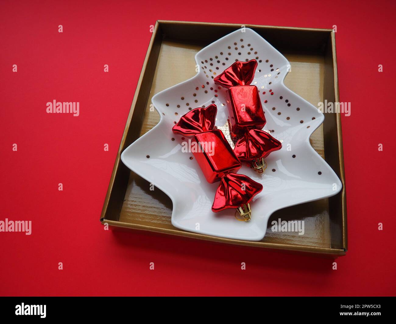 A plate in the shape of a Christmas tree with gold ornaments and a star in the middle. Gift wrapping. Red background. Candy in a red wrapper. Backdrop Stock Photo