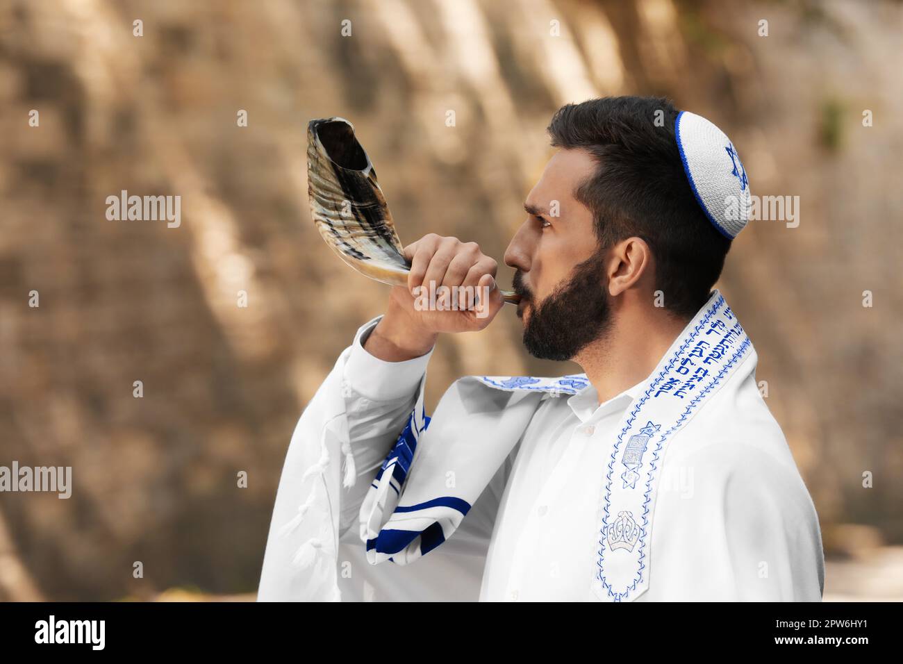 Jewish man blowing shofar on Rosh Hashanah outdoors. Wearing tallit with words Blessed Are You, Lord Our God, King Of The Universe, Who Has Sanctified Stock Photo