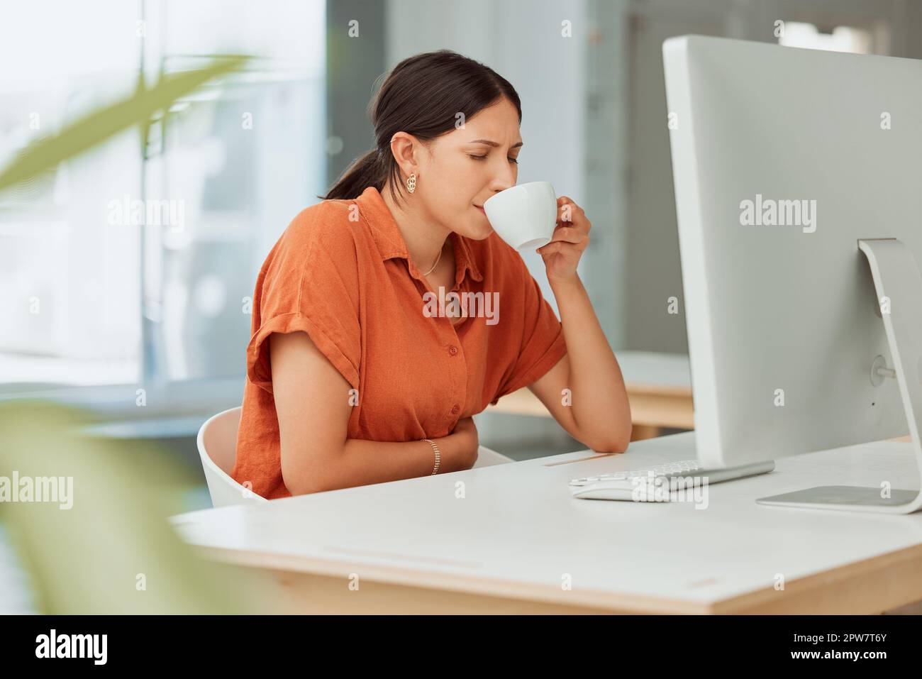 One hispanic business woman drinking warm tea as a remedy for sore tummy while feeling ill with menstrual stomach cramps in an office. Sick employee o Stock Photo