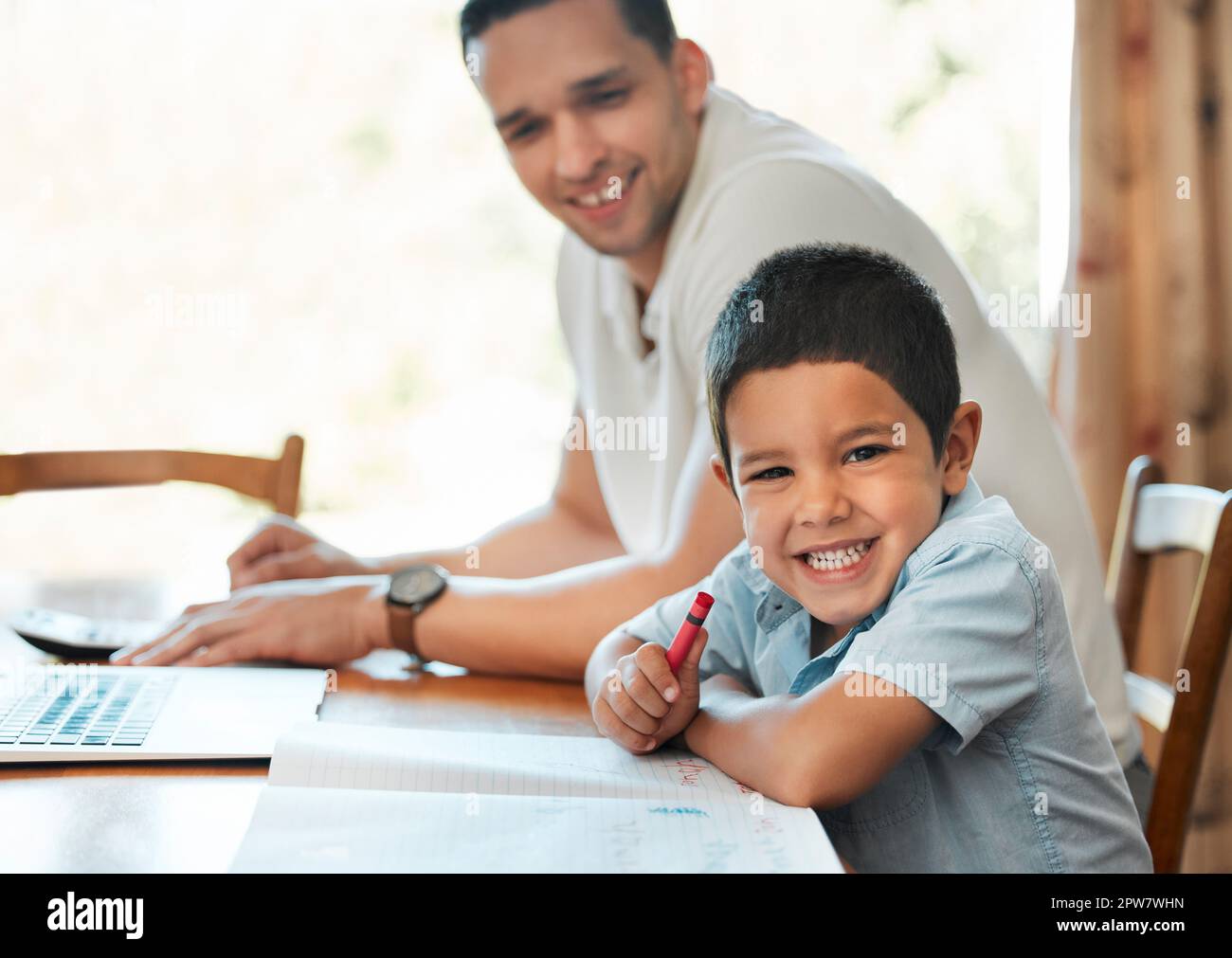 Excited preschooler boy drawing and writing with crayon at table at home while dad works remotely. Child sitting with parent during online education c Stock Photo