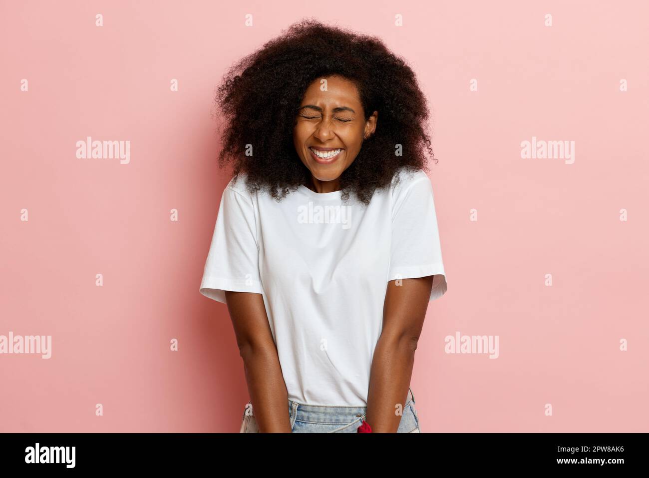 A portrait shot dark skinned curly haired shy and smiling woman in white t-shirt next to pink wall. High quality photo Stock Photo