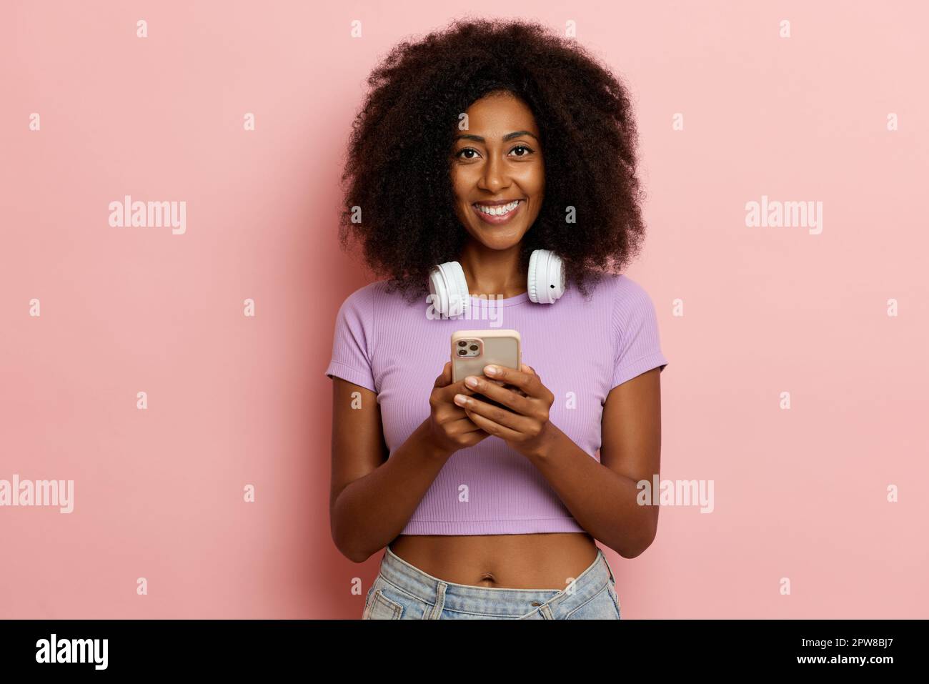 Attractive positive ethnic girl with fluffy hair, smiles toothily and looks to the camera, uses phone for messaging, dressed in casual cloth, poses ov Stock Photo