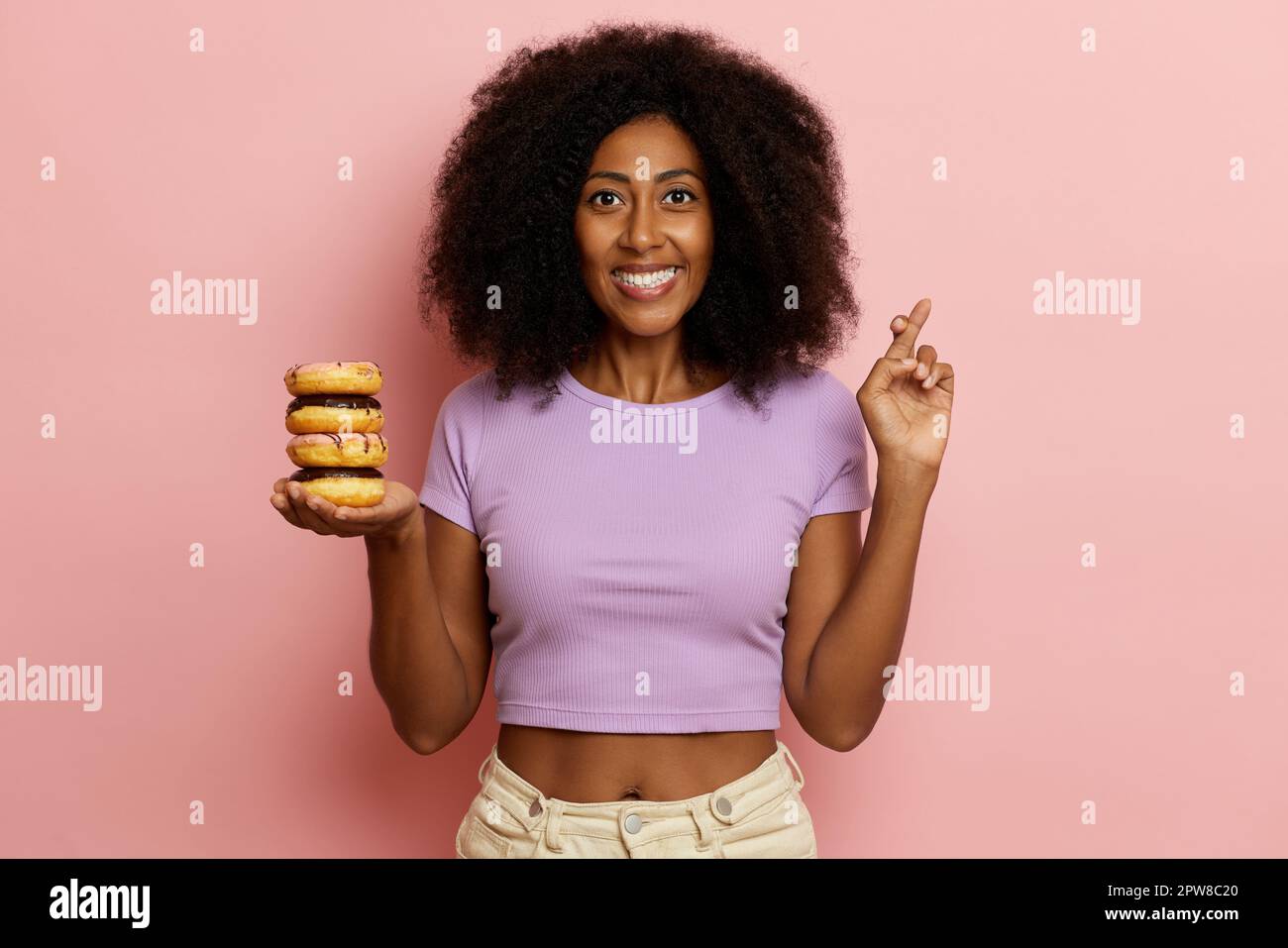 Hopeful curly haired brunette girl keeps delicious doughnuts, makes finger crossed gesture, makes a wish and smiles broadly, dressed in casual cloth, Stock Photo