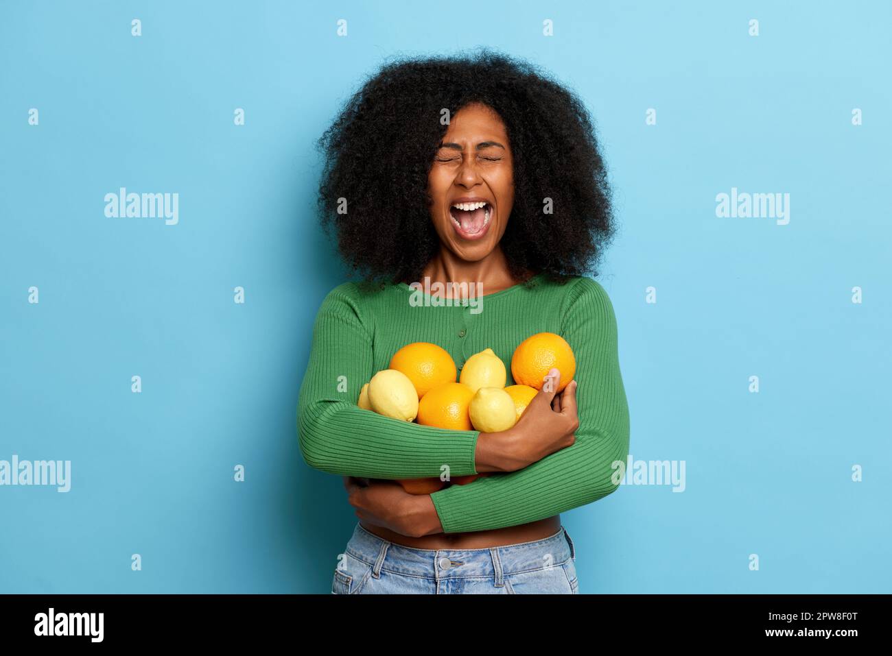 Indoor shot of emotional curly girl holding fresh oranges and lemons fruits, keeps mouth widely opened, dressed in casual cloth. High quality photo Stock Photo