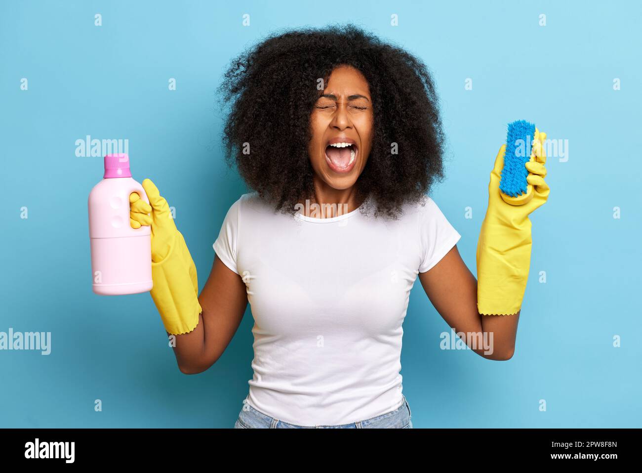 African curly haired woman screaming with brush and cleaning liquid in the hands, she wearing white t-shirt and yellow gloves. Stock Photo