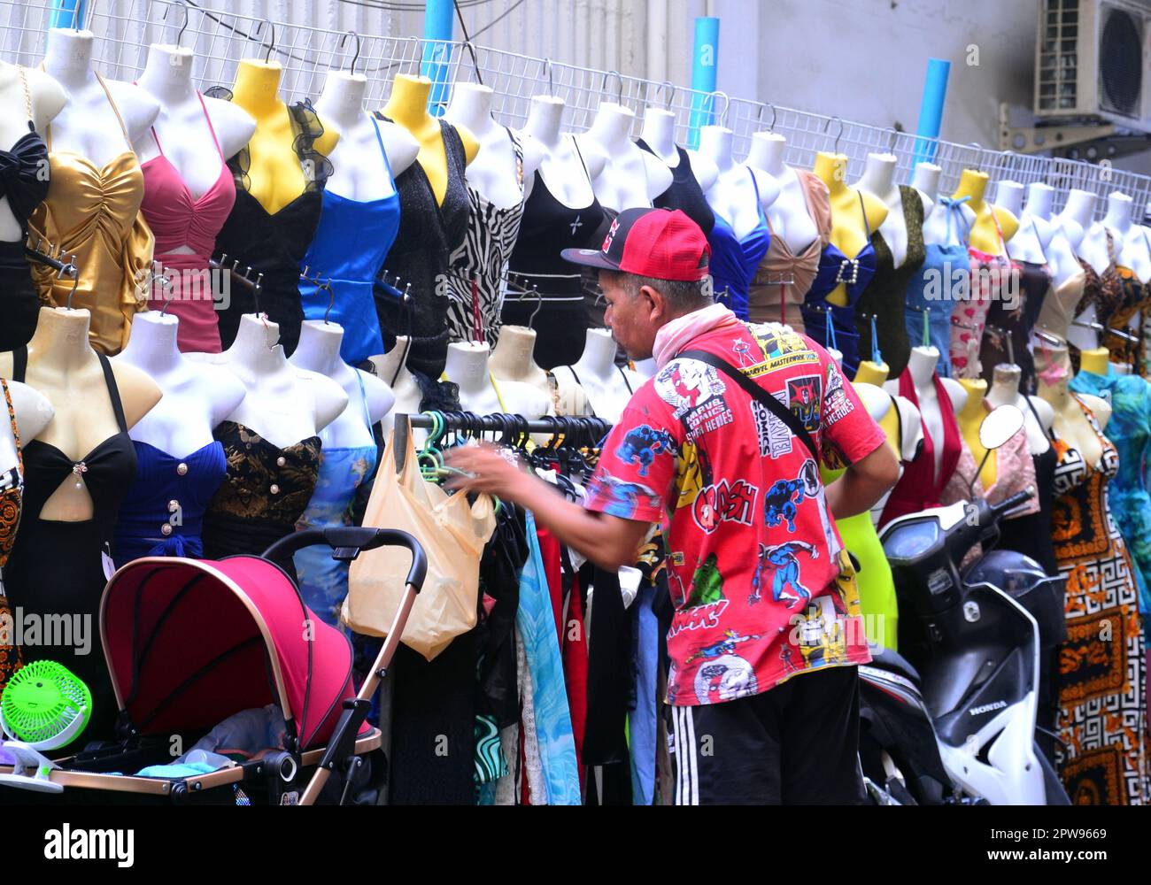 Working people, Bangkok, Thailand, Asia. A man prepares the display on a pavement clothes stall in Silom District. Baby in pram at bottom left: working people often take children to work when doing outside jobs in Thailand as they have no choice. A handy fence used to display the clothes. Work, worker, working, job, employment, earn money theme. Stock Photo