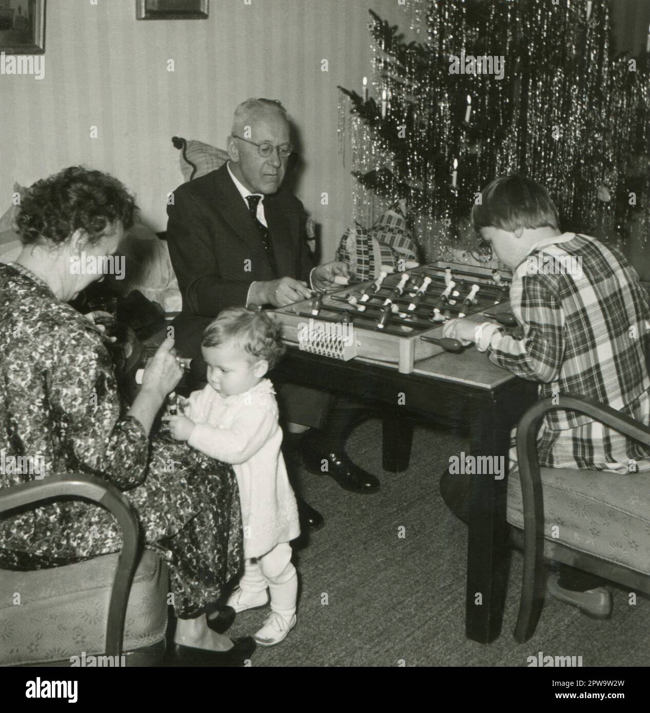Germany. 1960s. Grandparents with their grandchildren at Christmas. A grandfather is playing a table football game with his granddaughter, whilst a grandmother is paying attention to her infant grandchild. In the background is a Christmas tree decorated with lametta tinsel and lit candles. Stock Photo