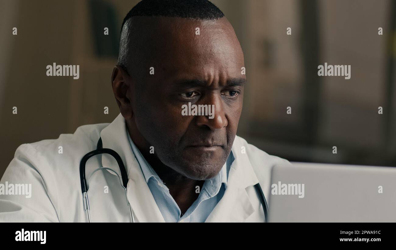Thoughtful wise experienced mature 50s male african american doctor medical professor general practitioner sit at hospital cabinet laboratory use Stock Photo