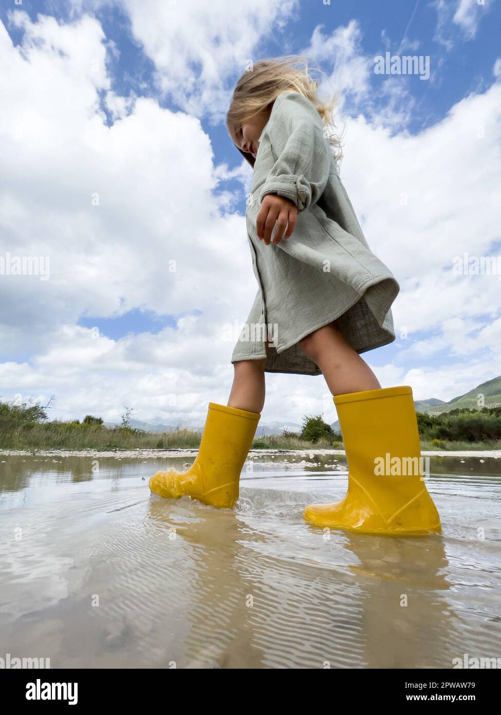 Little girl in yellow rubber boots walks through a puddle against a ...