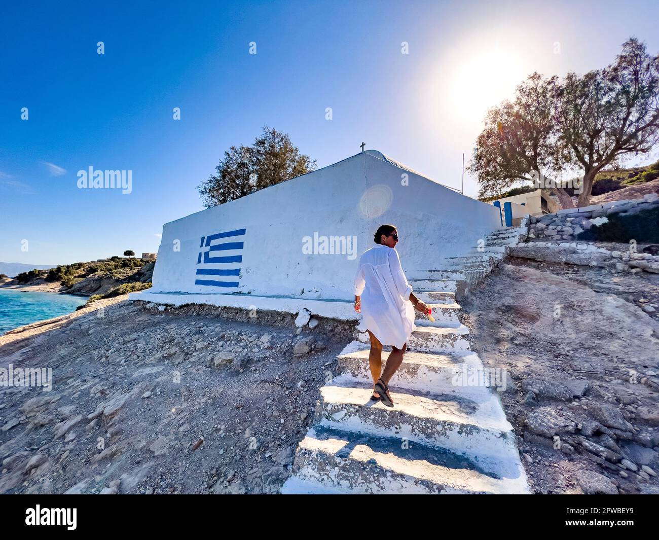 Female person walking up to a Greek orthodox church next to the sea. Travel destination concept. Place of worship for Christianity. Tourism background Stock Photo