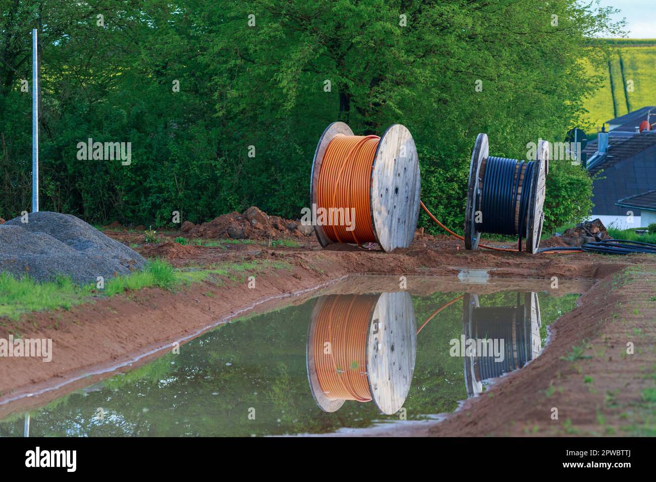 Glasfaserkabelrollen stehen an einer Straße und spiegeln sich in einem Erdloch voller Wasser Stock Photo