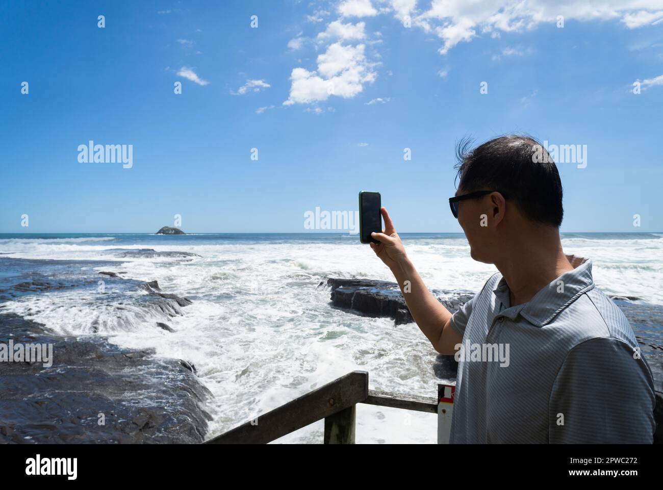 Tourist taking smartphone photos of big waves crashing onto rocks at Muriwai Beach. Auckland. Stock Photo