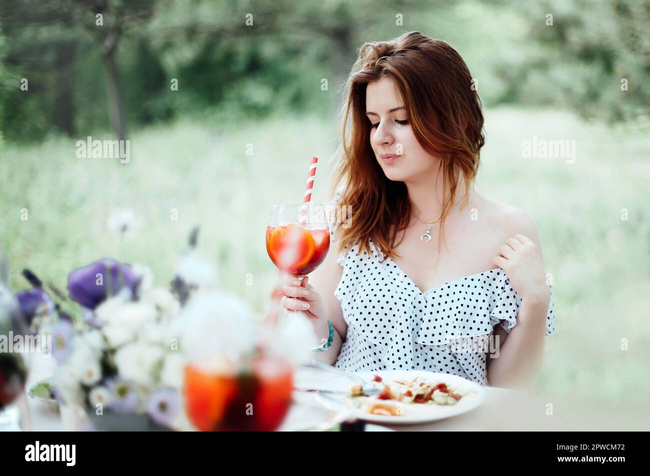 Happy young female with alcohol cocktail smiling and proposing toast while sitting at table during banquet in garden Stock Photo