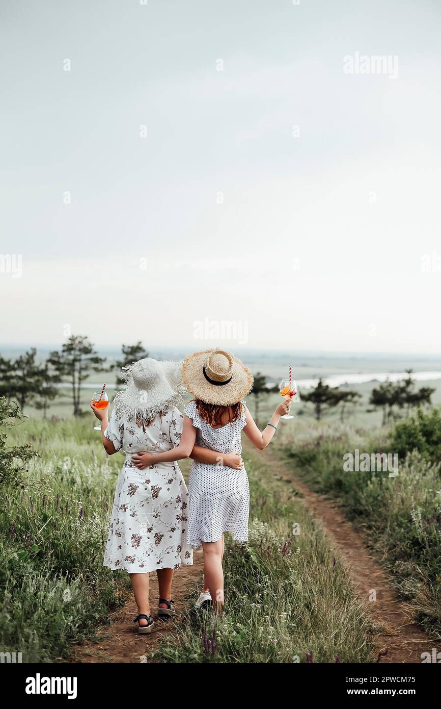 Back view of anonymous ladies with alcohol cocktails embracing each other and walking on path on summer day in nature Stock Photo