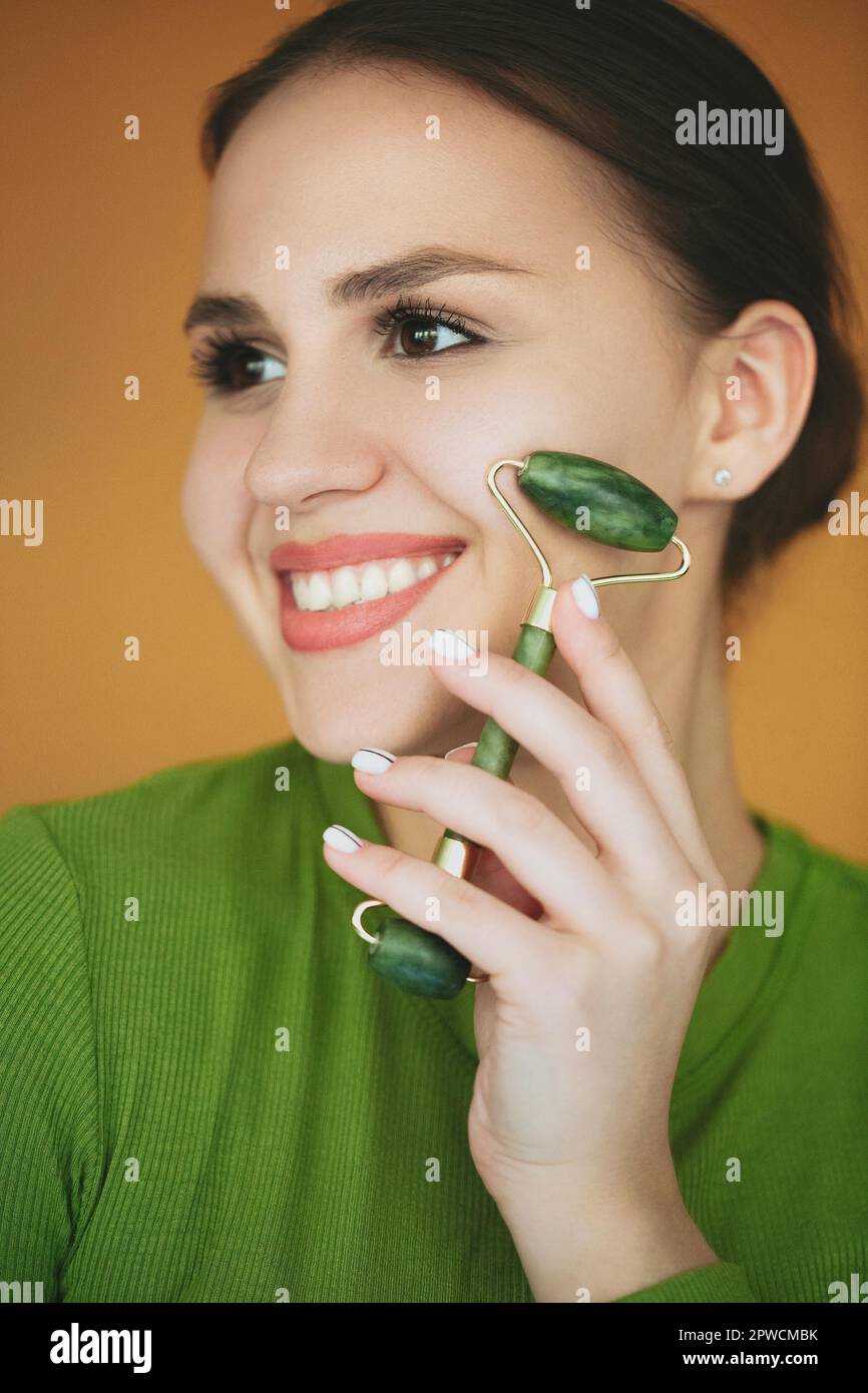 Smiling charming female massaging face with jade roller during skincare