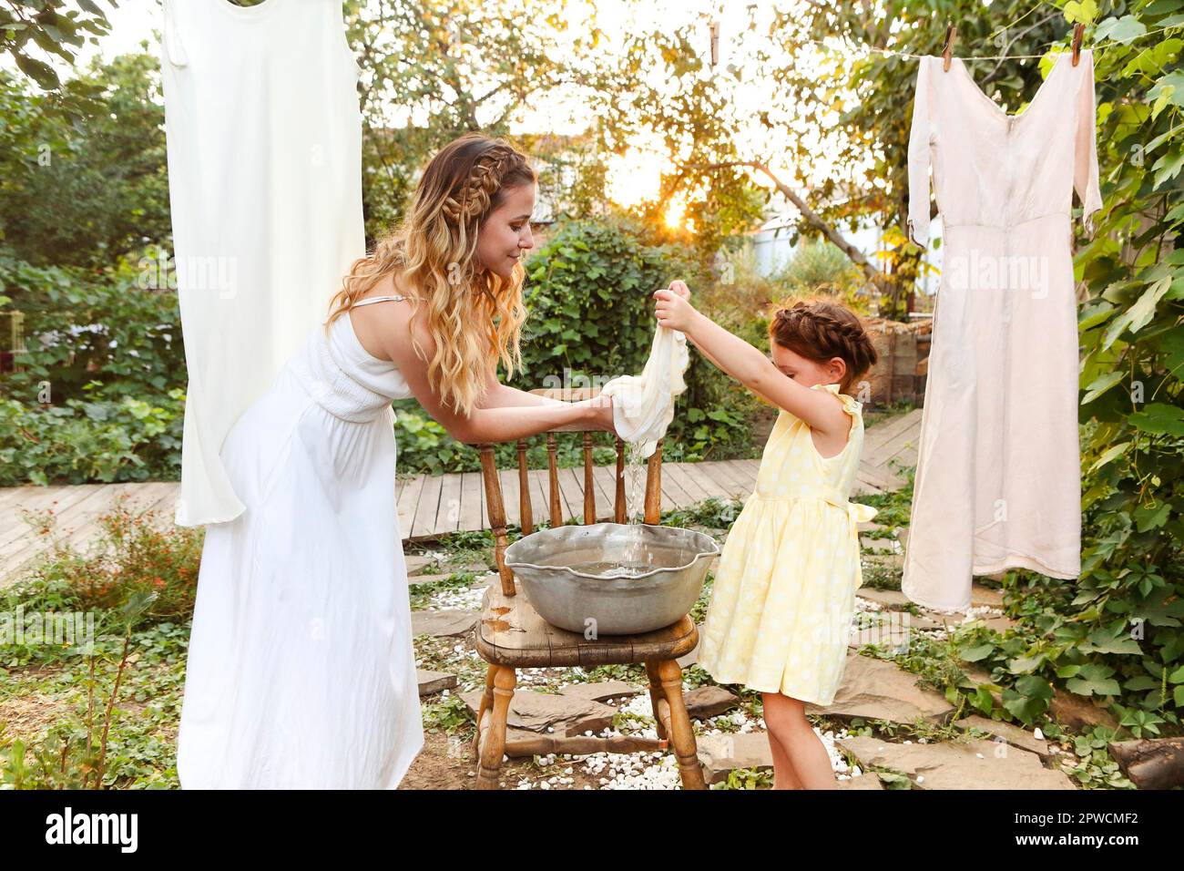 Charming little girl helping mother with wicker basket while doing chore and hanging laundry in backyard in summer evening Stock Photo