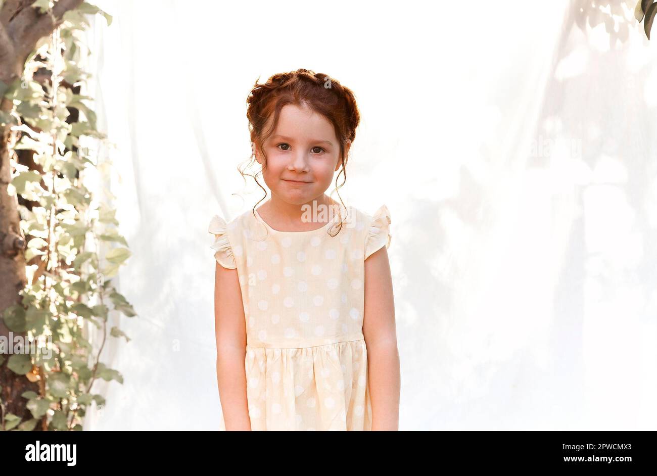 Close up portrait of cute little adorable redhead girl with curly hair with braid wearing yellow dotted dress looking at camera while playing in Stock Photo