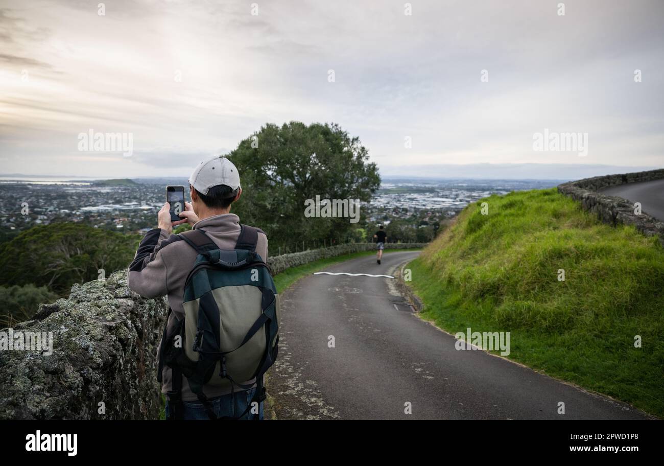 Tourist taking photos with smartphone at One Tree hill. Out-of-focus people running. Auckland. Stock Photo