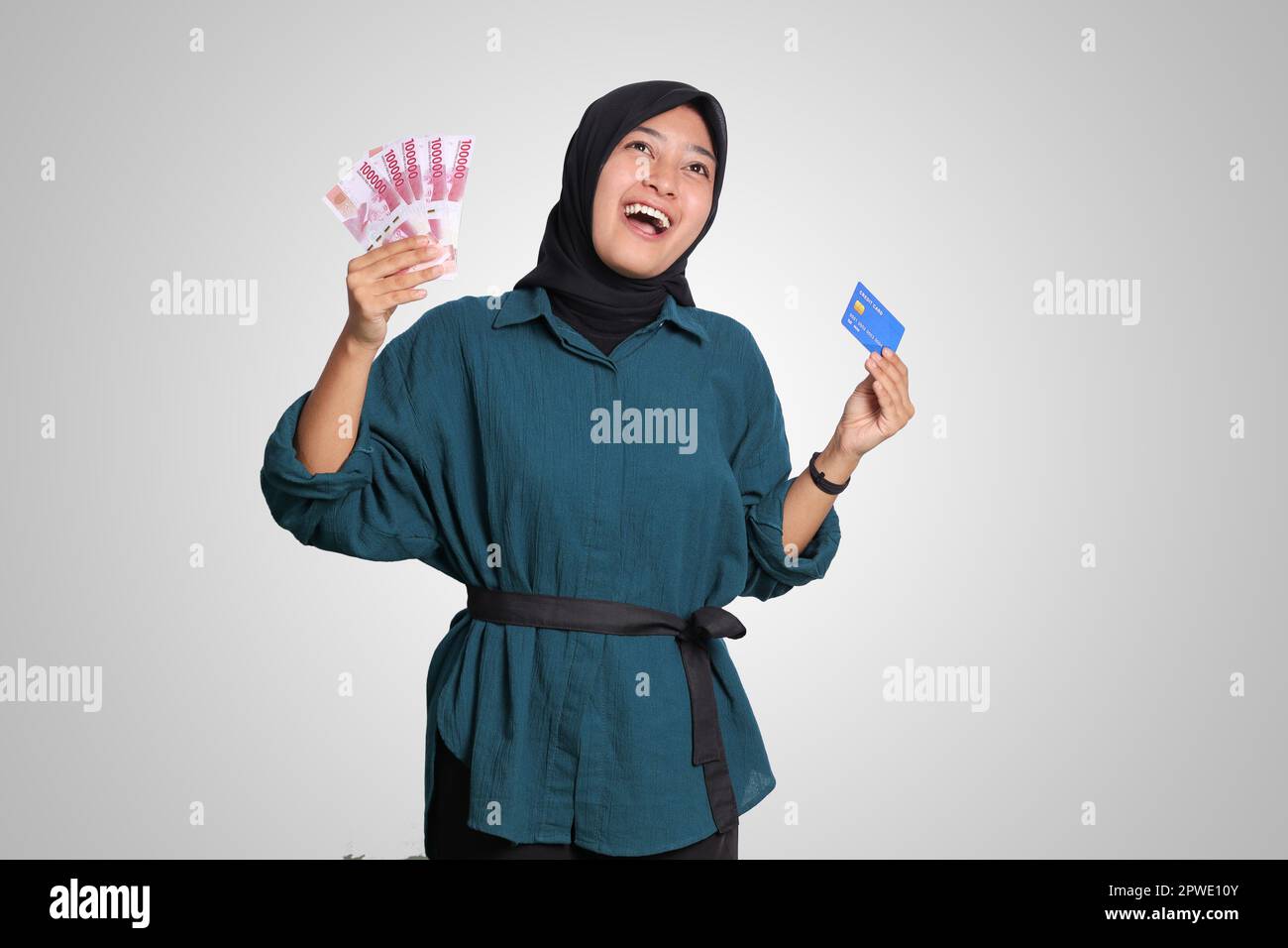 Portrait of cheerful Asian muslim woman with hijab, showing one hundred thousand rupiah while holding a credit card. Financial and savings concept. Is Stock Photo