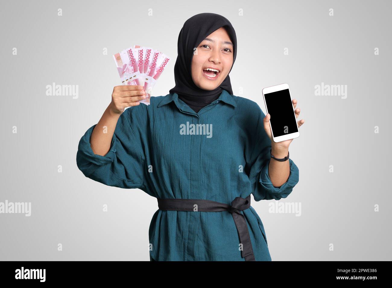 Portrait of excited Asian muslim woman with hijab, showing one hundred thousand rupiah while showing blank screen mockup mobile phone. Financial and s Stock Photo