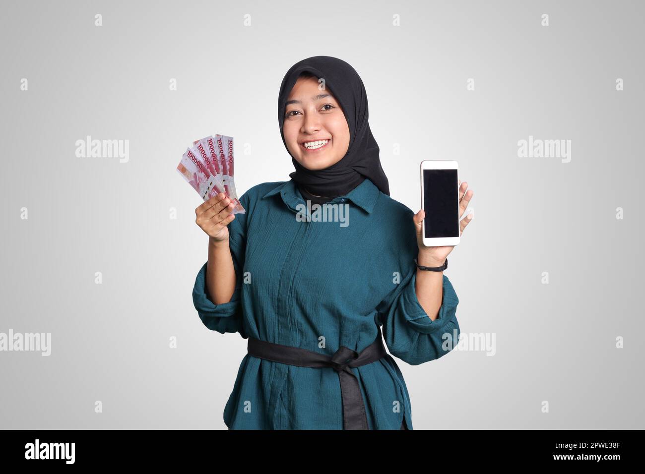 Portrait of excited Asian muslim woman with hijab, showing one hundred thousand rupiah while showing blank screen mockup mobile phone. Financial and s Stock Photo
