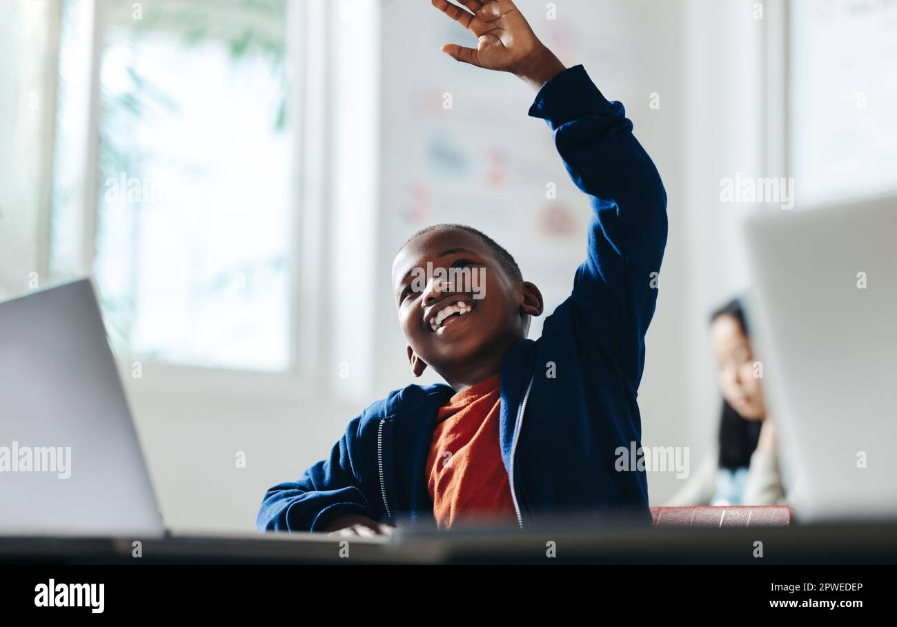Happy young boy raising his hand to answer a question in a digital literacy classroom. Sitting with a laptop in front of him, the primary school stude Stock Photo
