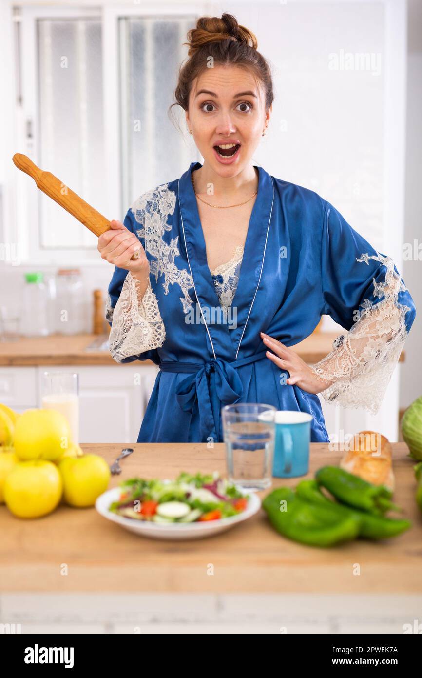 Angry woman screaming and waving rolling pin in home kitchen Stock Photo