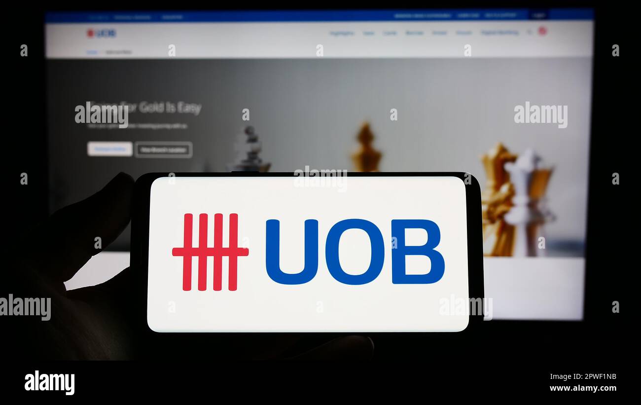 Person holding smartphone with logo of company United Overseas Bank Limited (UOB) on screen in front of website. Focus on phone display. Stock Photo