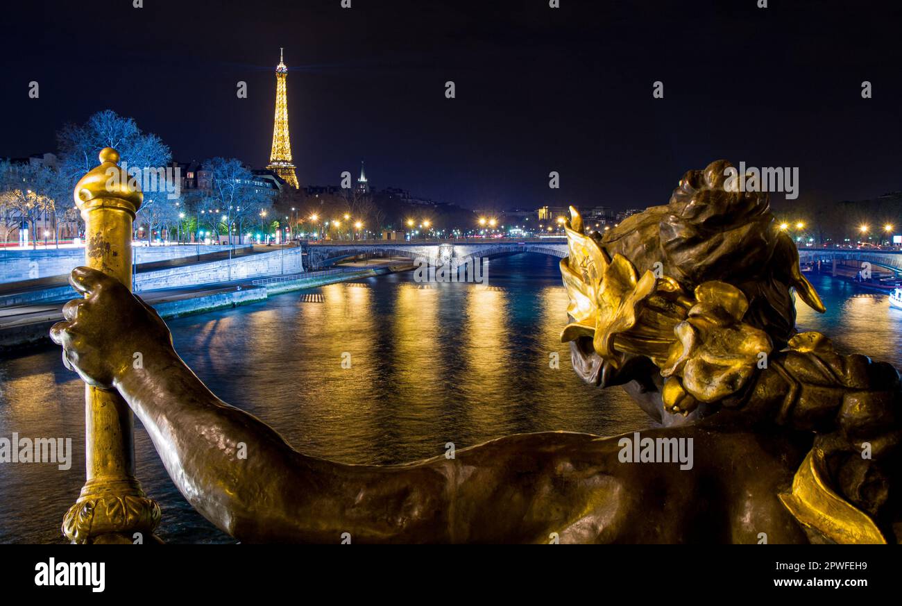 View of the Seine river by night with the Eiffel Tower in the background, as seen from behind a golden nymph statue on the Alexander III bridge in Par Stock Photo