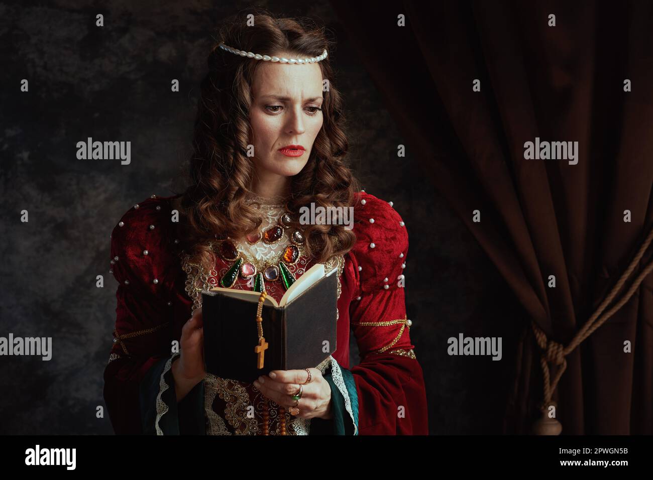pensive medieval queen in red dress with book and rosary on dark gray ...