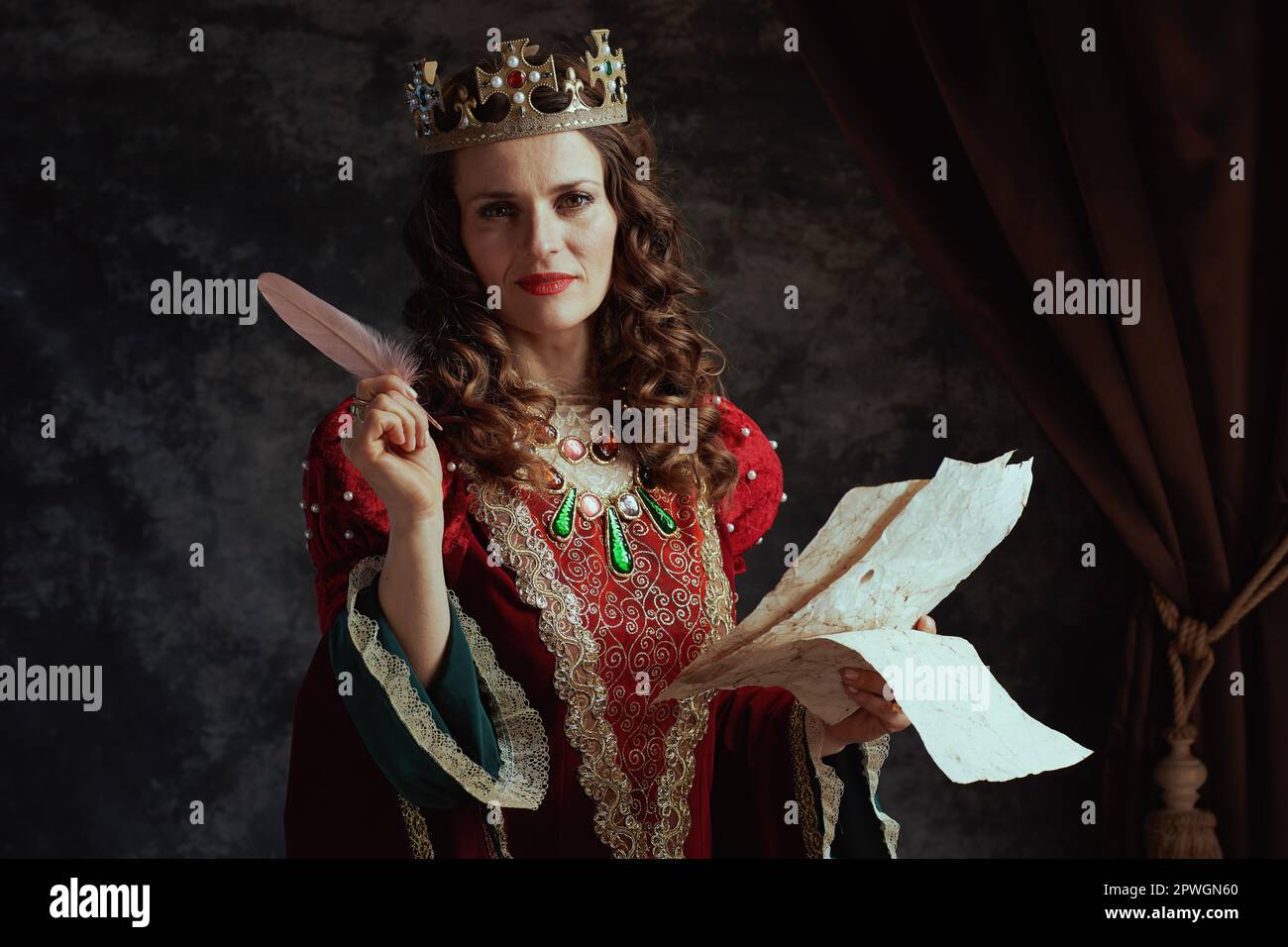 happy medieval queen in red dress with parchment and crown on dark gray background. Stock Photo