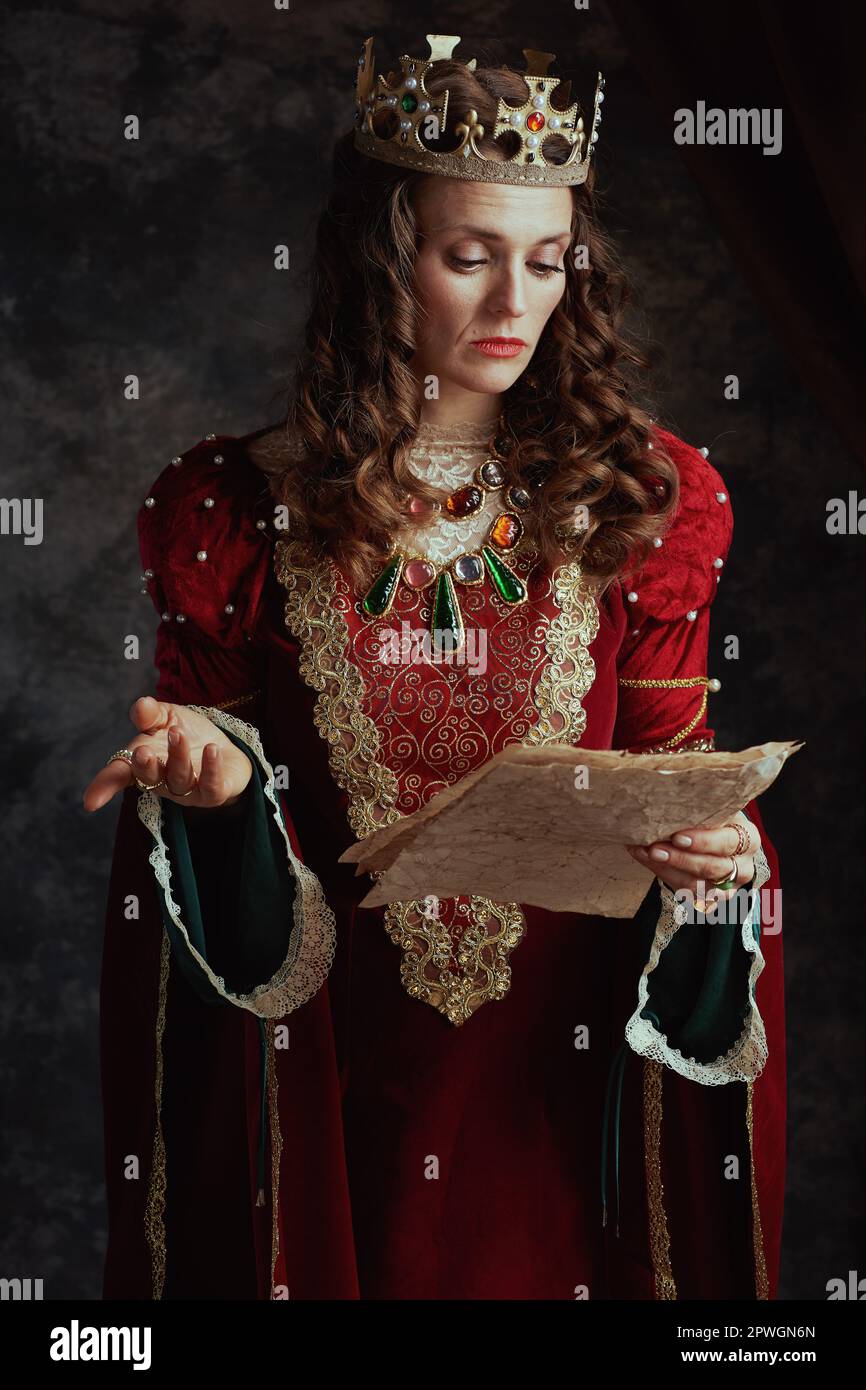 medieval queen in red dress with parchment and crown on dark gray background. Stock Photo