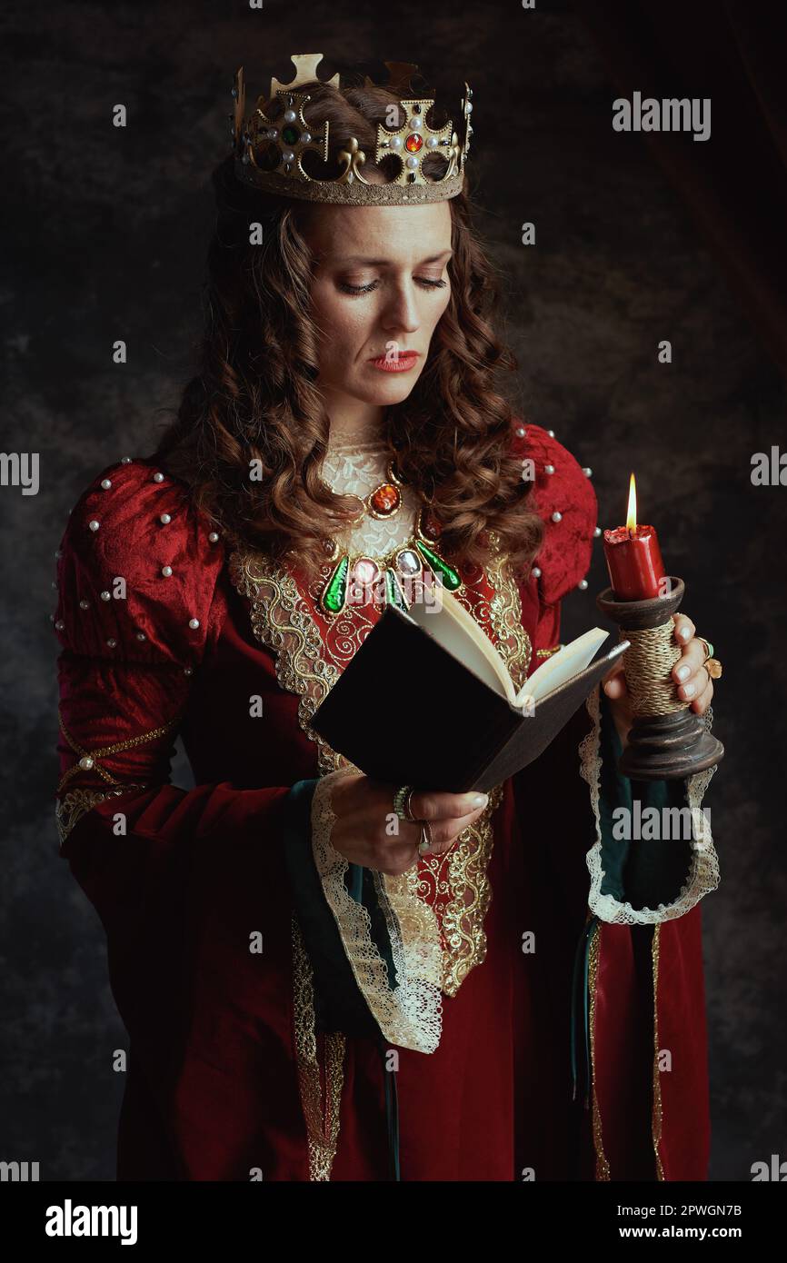 medieval queen in red dress with book, candle and crown on dark gray ...