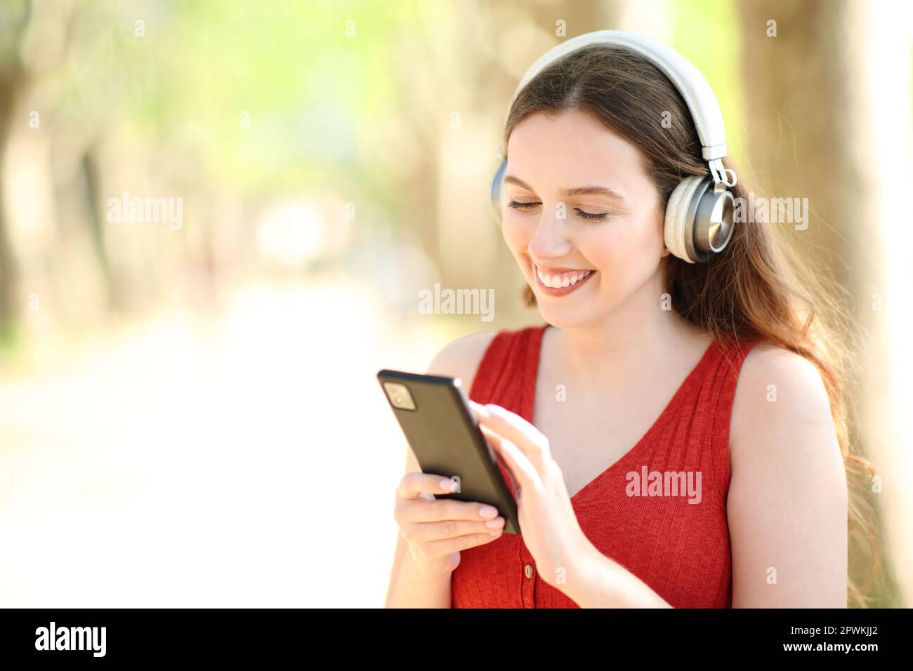 Happy woman wearing headphone listening to music using smart phone standing in a park Stock Photo