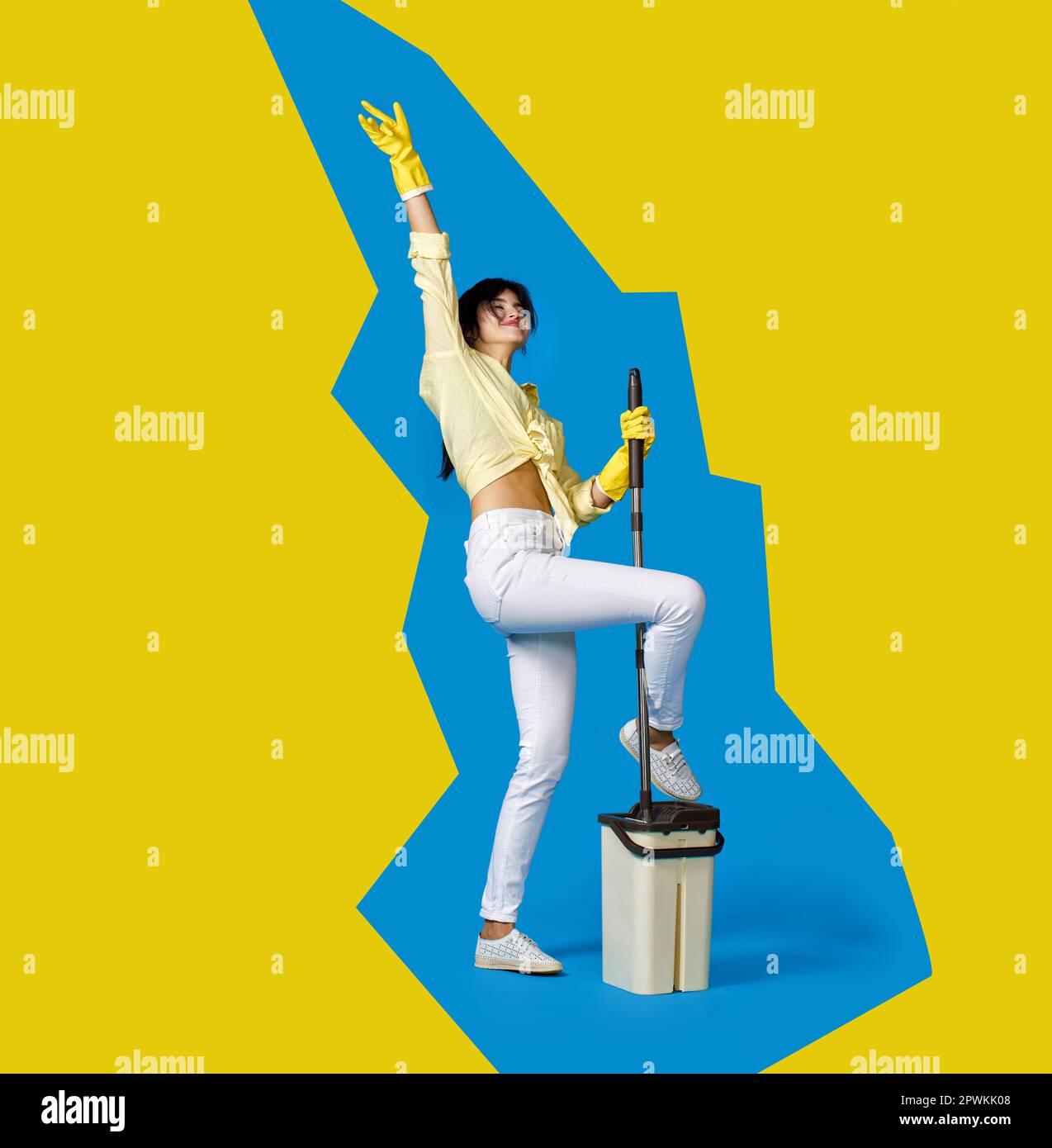 funny housewife girl in rubber gloves washing floor with mop and dancing on blue and yellow background. Full length Stock Photo
